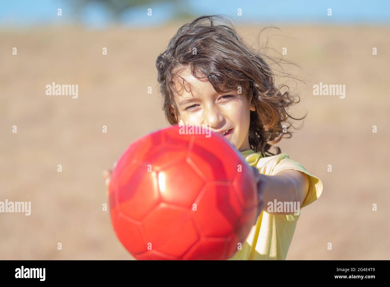 little boy with long hair and yellow t-shirt plays with his red ball in a field on a sunny summer day Stock Photo