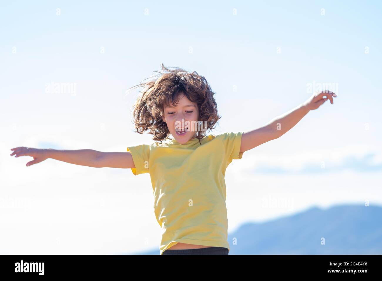 smiling boy with long hair jumps and has fun playing with open arms. blue sky background on a sunny day Stock Photo