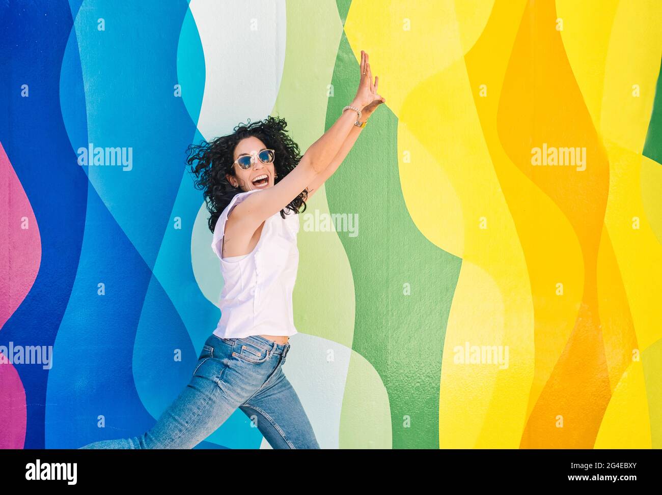 Young brunette woman with curly hair, jeans and sunglasses has fun while jumping on a colorful wall. leisure and free time concept Stock Photo