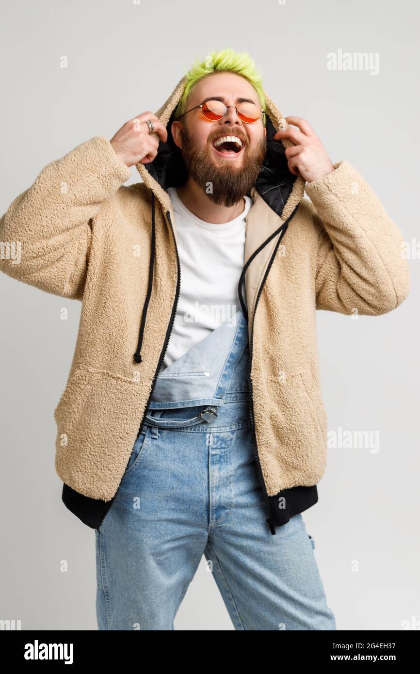 Extremely happy bearded young adult man wearing denim overalls and jacket, laughing out loud, keeping hands on his hood, hears funny joke. Indoor stud Stock Photo