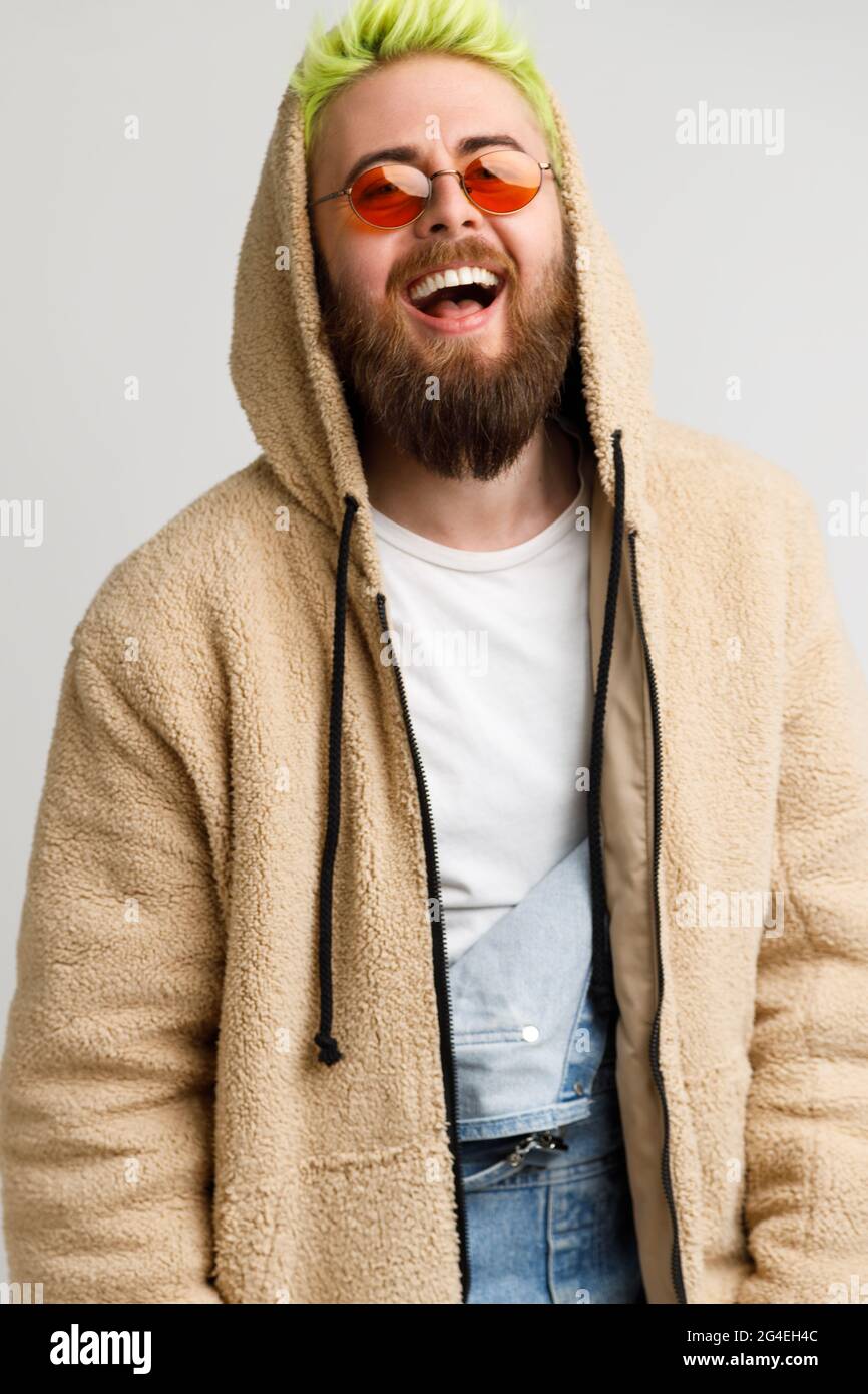 Handsome young adult bearded model man wearing beige jacket looking at camera and laughing out loud, expressing positive and optimism. Indoor studio s Stock Photo