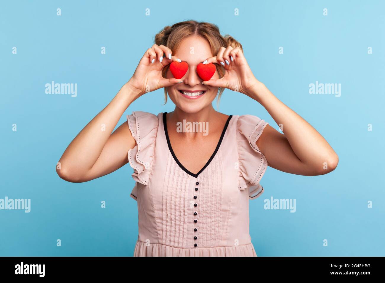 Portrait of beautiful blonde haired female with hair buns in dress covering eyes with heart shape symbols, expressing romantic feelings, friendship. I Stock Photo