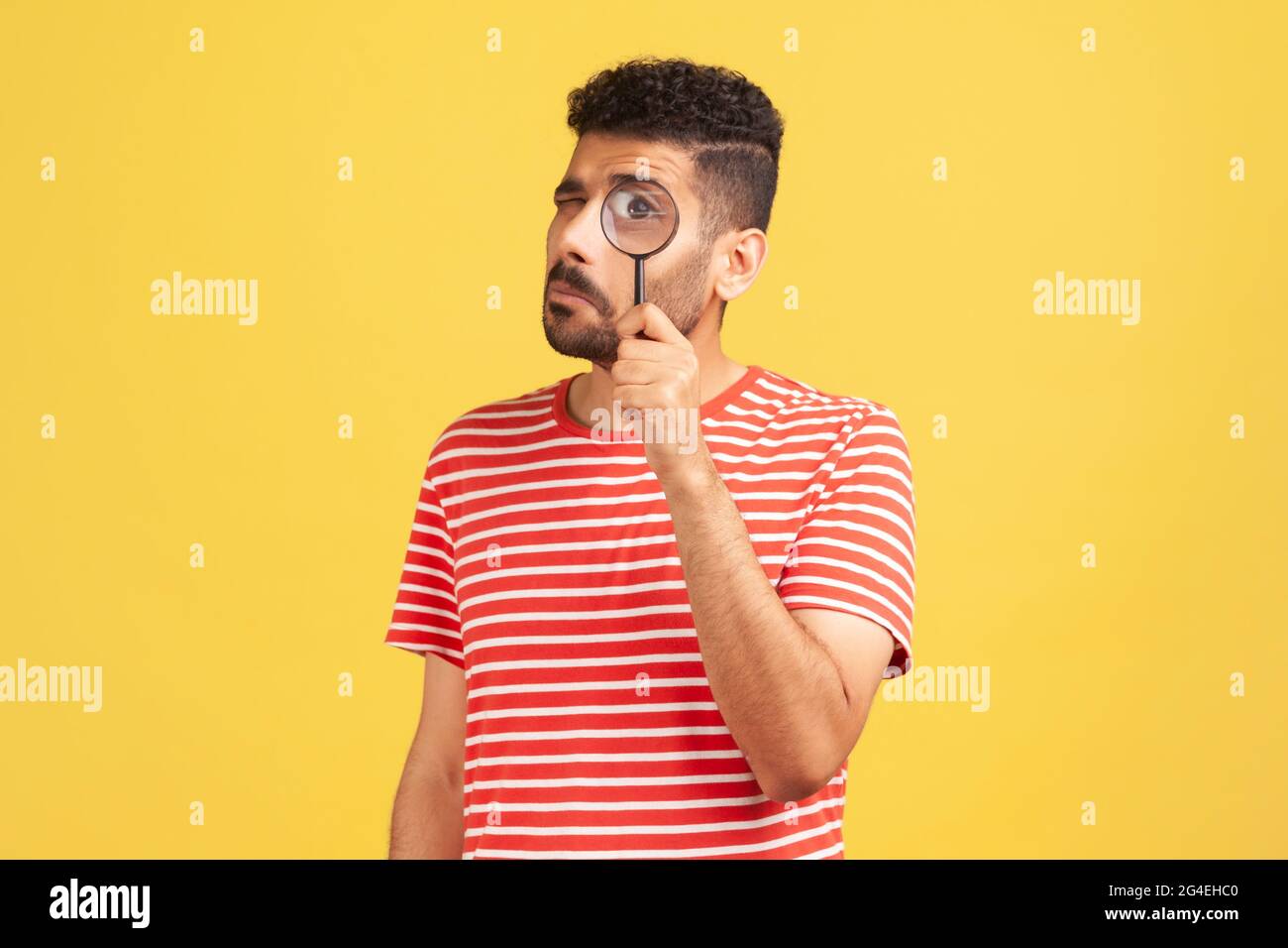 Serious bearded man in red striped t-shirt standing, holding magnifying glass and looking at camera with big zoom eye, verifying authenticity Stock Photo