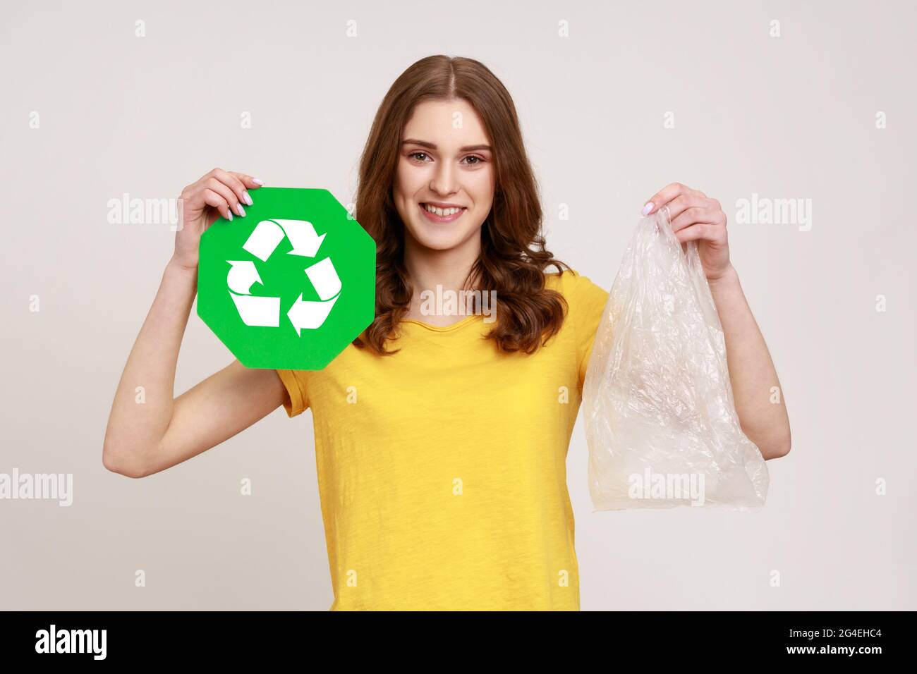 Portrait of beautiful happy teenager girl with brown hair wearing yellow casual T-shirt, holding in hands plastic package and green recycle sign. Indo Stock Photo