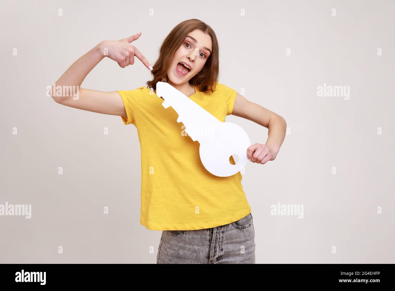 Portrait of funny positive teenager girl in casual style yellow T-shirt pointing at big paper key and looking at camera with toothy smile, purchase. I Stock Photo