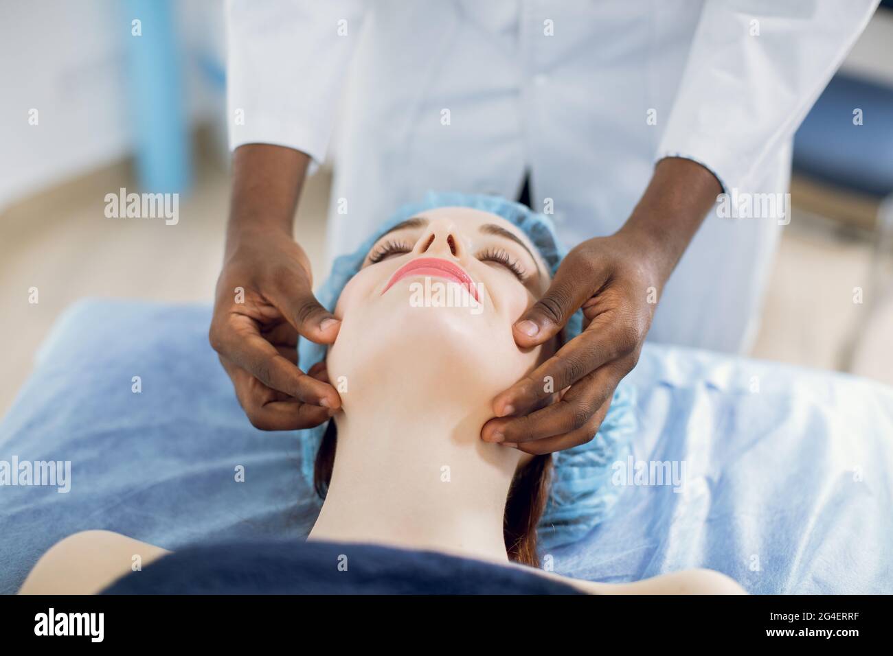 Relaxing treatment in medicine. Caucasian woman in disposable cap, lying on the couch, enjoying facial massage with closed eyes in spa salon or clinic. Hands of male black chiropractor doing massage Stock Photo