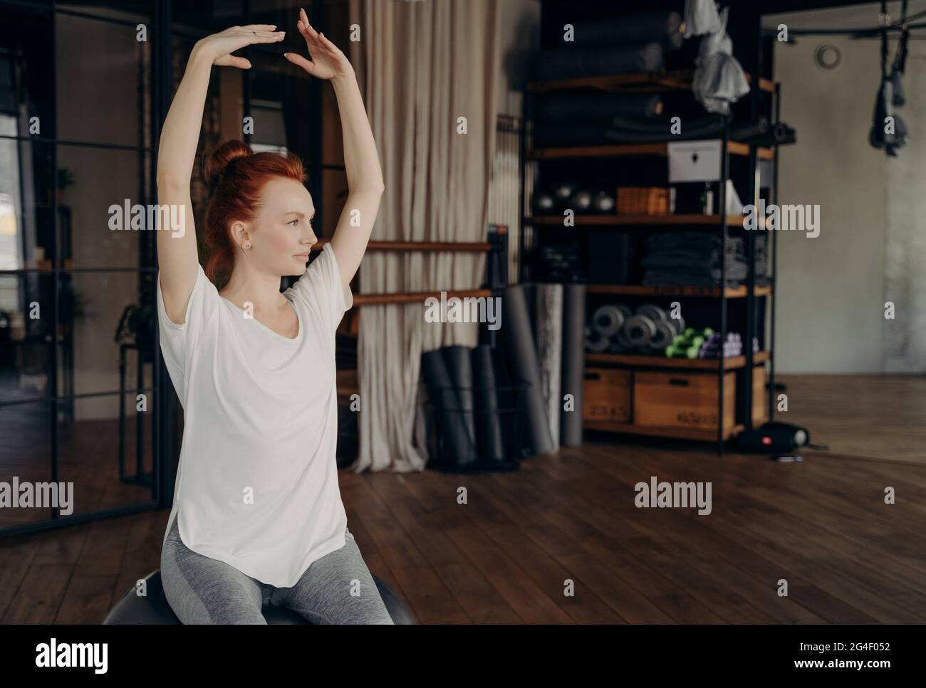 Sportive young woman with red hair sits on big pilates ball with hands lifted over head Stock Photo