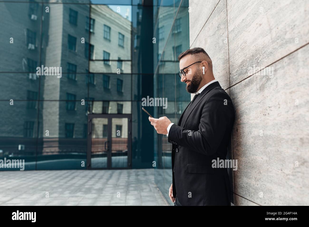 Entrepreneur listening to music in wireless earphones and using smartphone, standing outdoors, empty space, side view Stock Photo