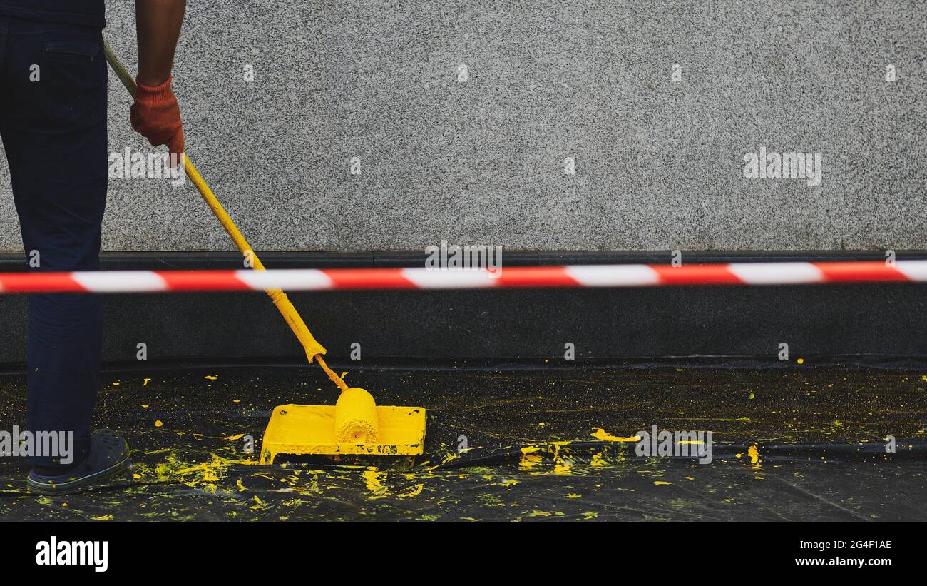 Signal tape beside painting the building. Man painting a wall in yellow colour. Paint roller in a paint tray near a grey wall. Black plastic cover on Stock Photo