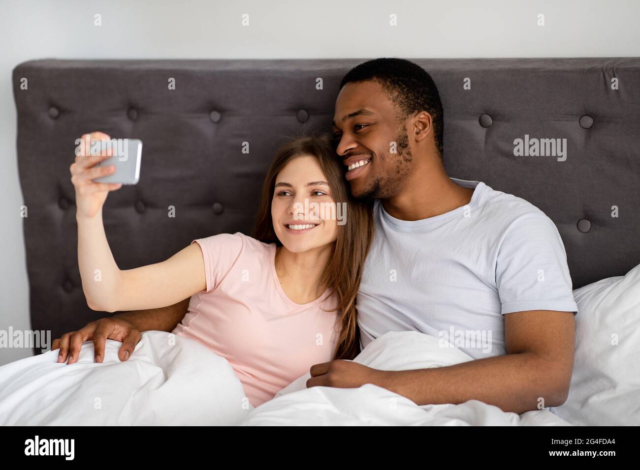 Millennial Multiracial Couple With Smartphone Taking Selfie In Bed Before Going To Sleep