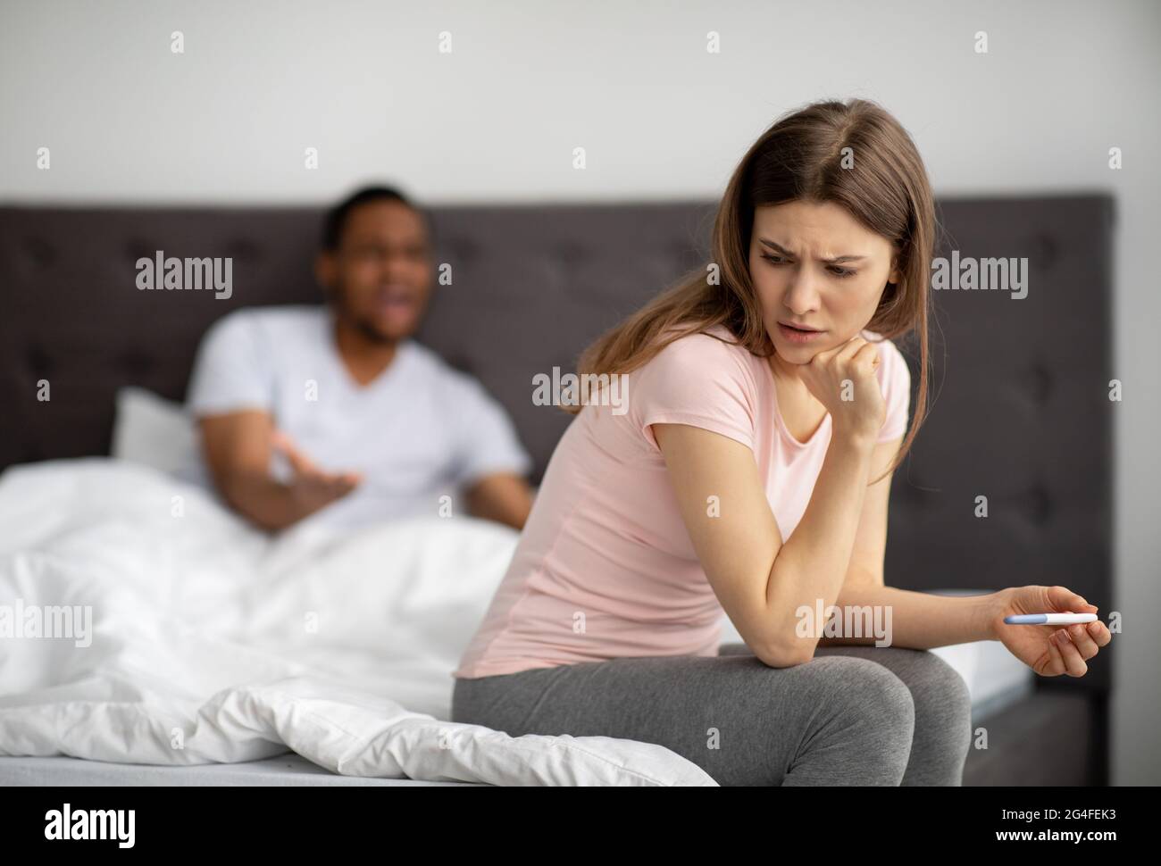 Unhappy young woman sitting on bed with pregnancy test, black husband screaming at her, indoors Stock Photo