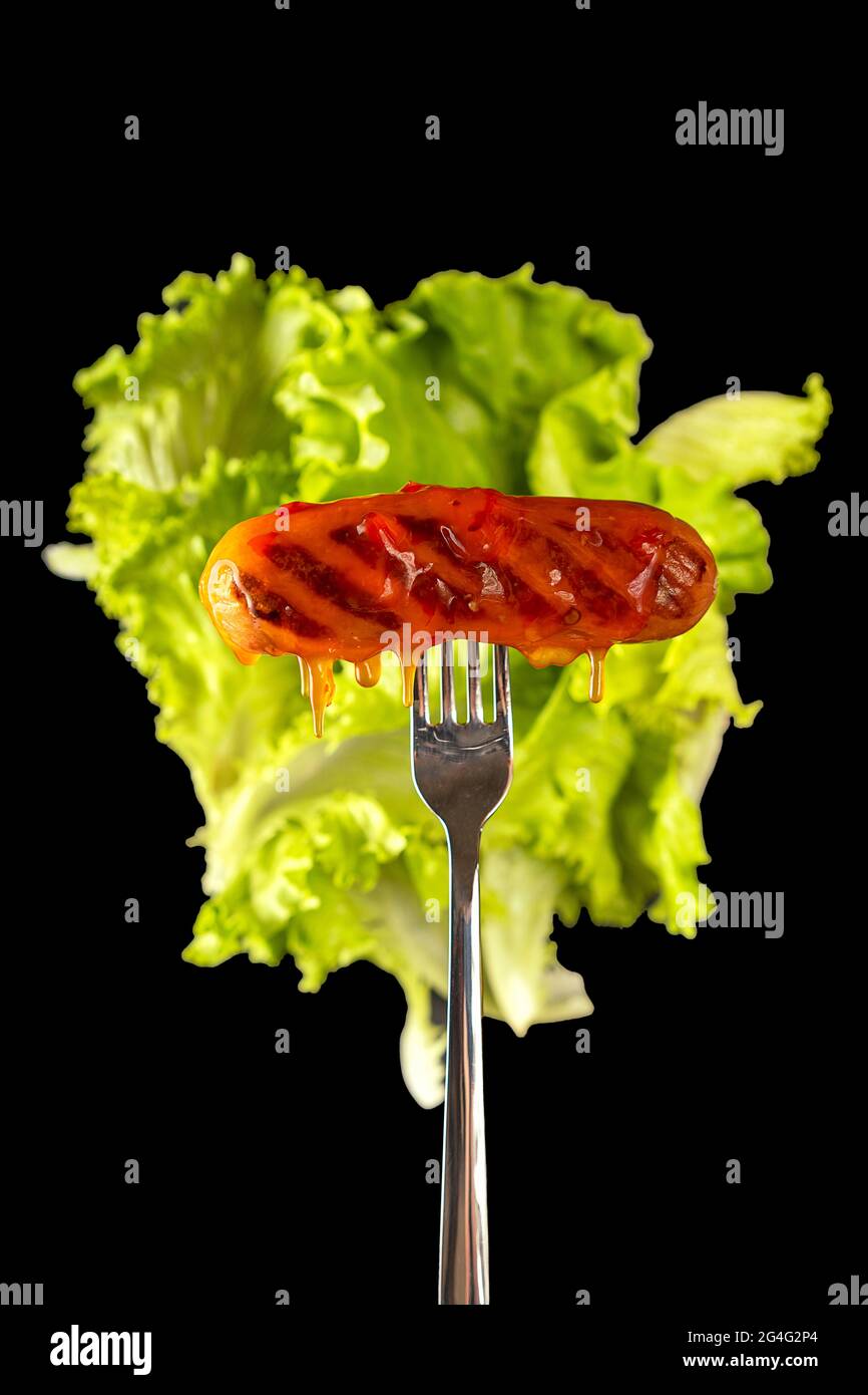 Grilled sausage on a fork, lettuce leaves in the background. Isolated on a black background. Cholesterol, junk food. Selective focus. Stock Photo