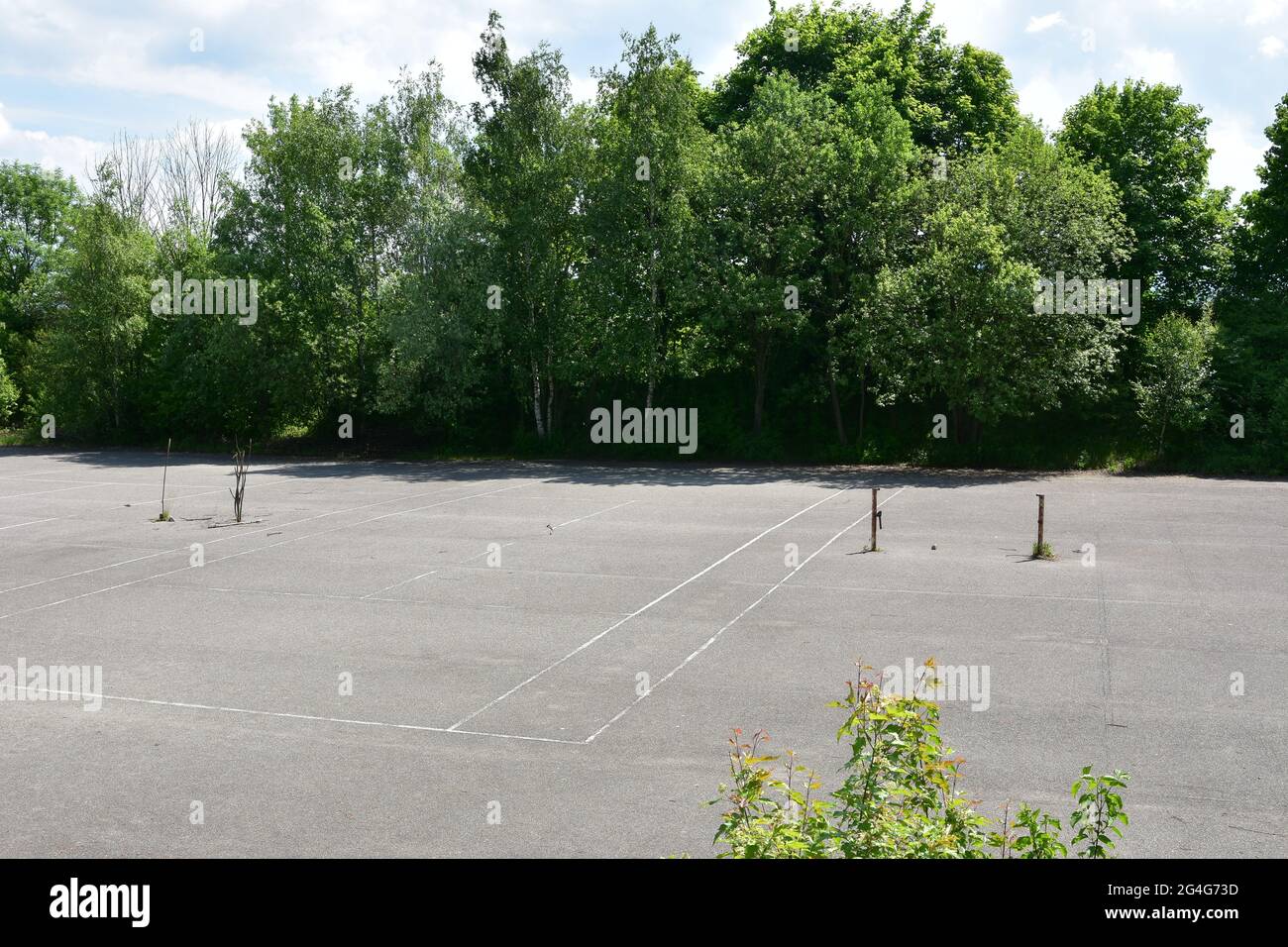 Old abandoned, desolate concrete hard tennis courts surrounded by trees and bushes under blue sky. Rusty tennis net posts are fixed to the asphalt . Stock Photo