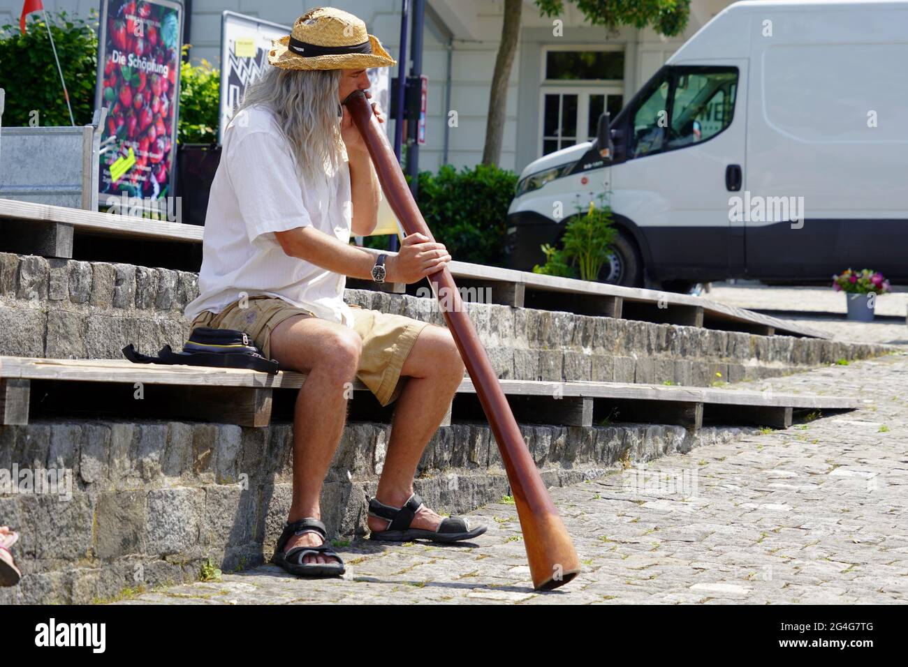 Man playing a didgeridoo in city Zug in Switzerland. He has long grey hair, is wearing straw hat, white shirt, shorts and sandals. Stock Photo