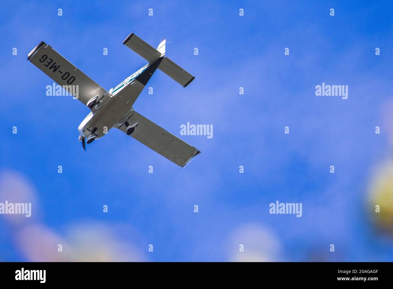 A portrait of a small sports airplane flying through a blue sky, with some small tin clouds behind the plane and some blurry leaves in the foreground. Stock Photo