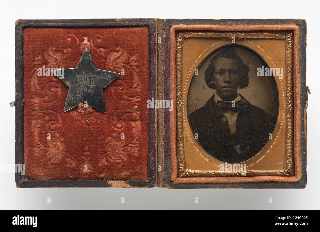 Tintype of Creed Miller, an African American soldier belonging to the Kentucky 107th Regiment, Company E (and later Company C). The tintype is encased in a copper scrolled frame with an oval window within a red velvet-lined, brown leather box. The tintype depicts Creed from the waist up in a dark overcoat, white collared shirt and bow-tie. His hair is styled and parted to the side. His miltary identification pin is fastened to the red velvet fabric on the interior of the case. The pin is a silver five pointed star with &quot;C. MILLER/ Co. E/ 107/ USCT&quot; engraved at center. The front and b Stock Photo