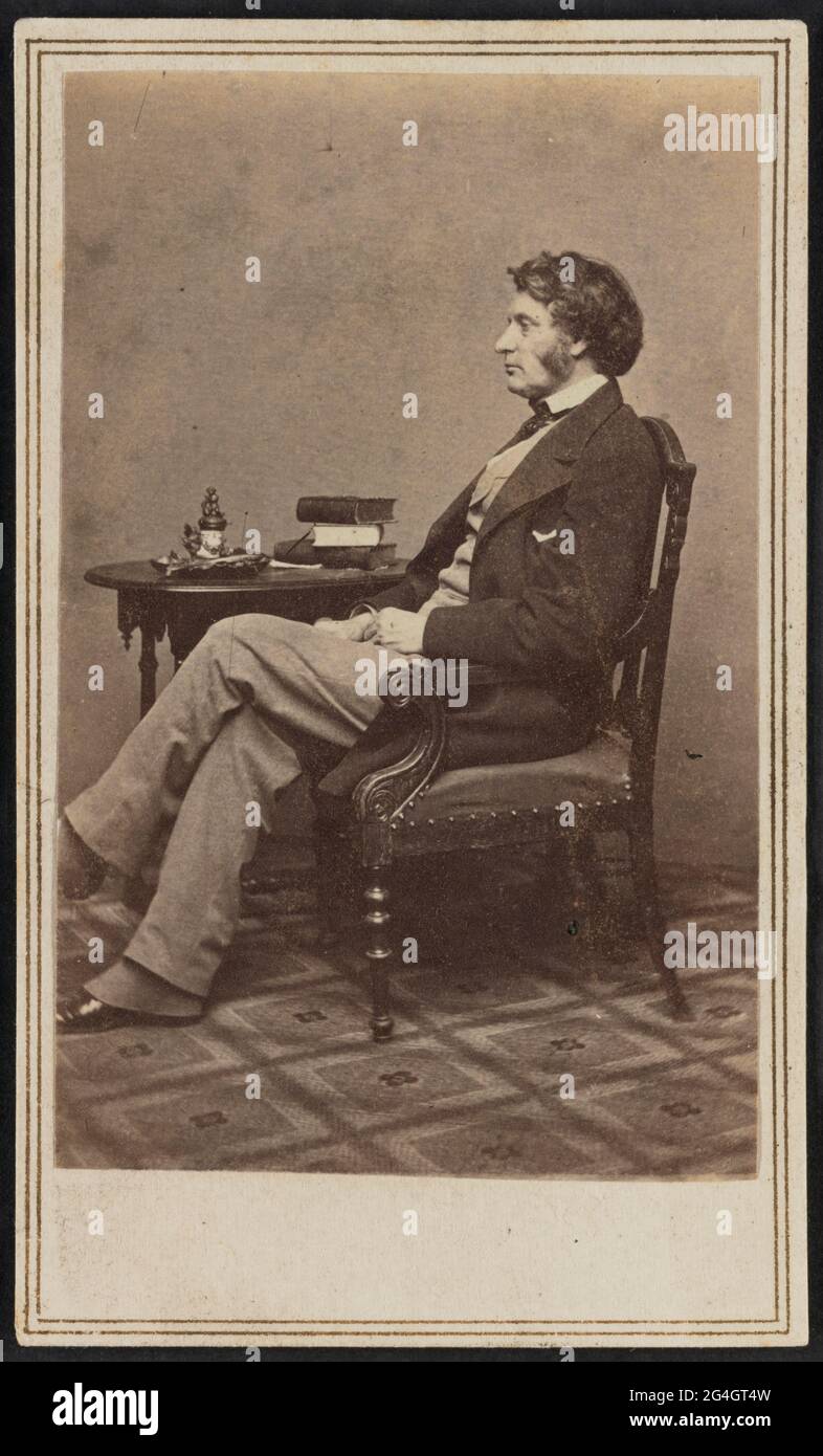 Carte-de-visite of American abolitionist Charles Sumner in full length seated profile. Sumner is pictured with his left profile facing the camera. His hands are resting in his lap and his left leg is crossed over his right leg at the knee. He is wearing a light colored vest and trousers with a white shirt, dark tie, and dark jacket. Spats peak out below his pant legs, partially covering his dark colored low-heeled shoes. His hair is moderately long and he has long sideburns. Sumner is seated in a carved armchair with an upholstered seat with a round wooden side table behind him. A stack of boo Stock Photo
