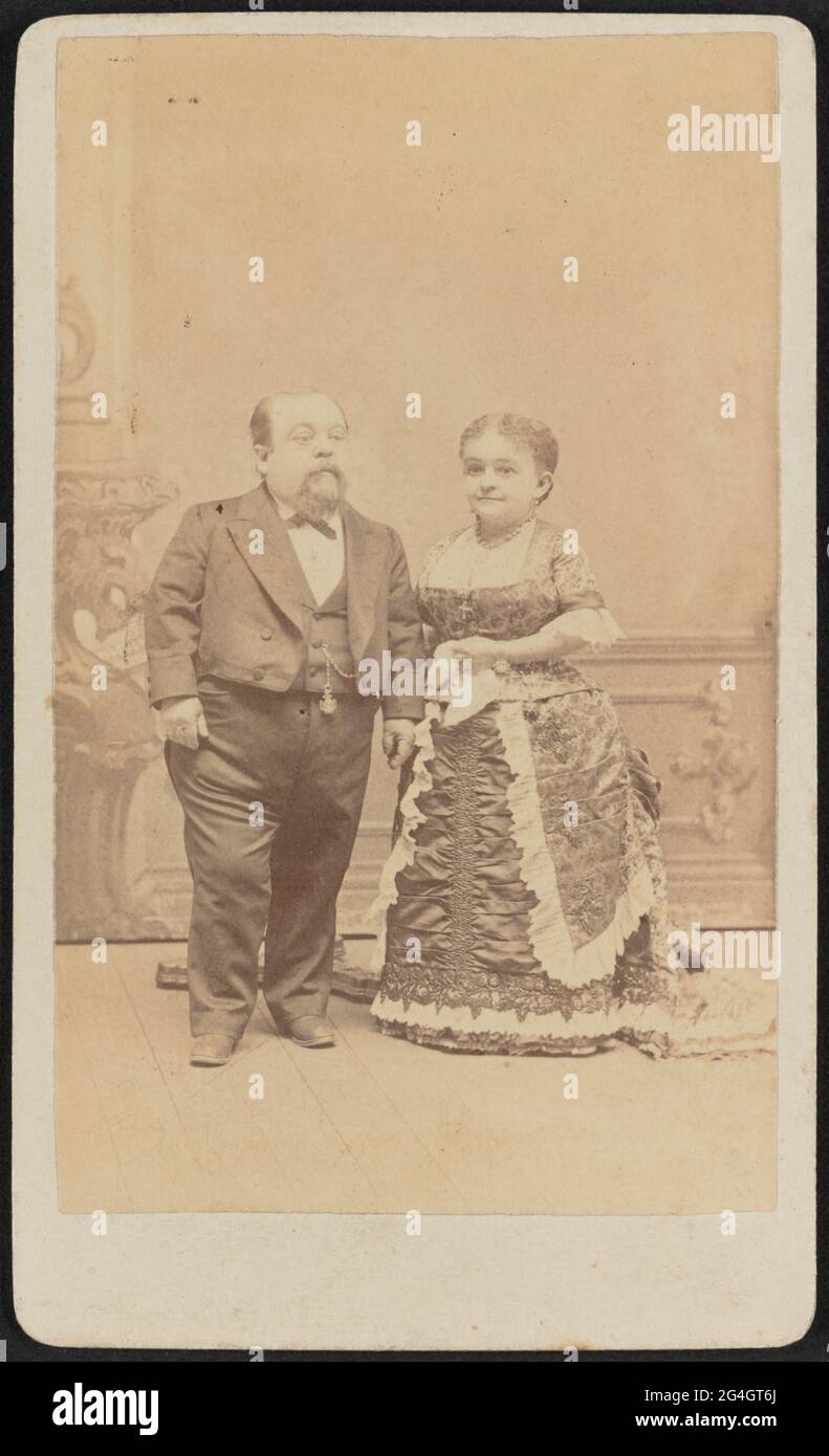 Carte-de-visite of Tom Thumb and Lavinia Warren shown in full portrait. Both are standing and facing slightly toward each other looking off frame. Thumb is on the left facing side of the image and wears a dark three-piece suit, white shirt, dark bowtie, and has a pocket watch on a chain hanging from his vest button. He is balding and has a mustache and goatee. Warren wears a dark colored floral damask evening dress with a bustle and white lace trimming on her cuffs and skirt. She has on drop earrings and wears a short beaded necklace and a longer chain necklace with a cross at the end that han Stock Photo