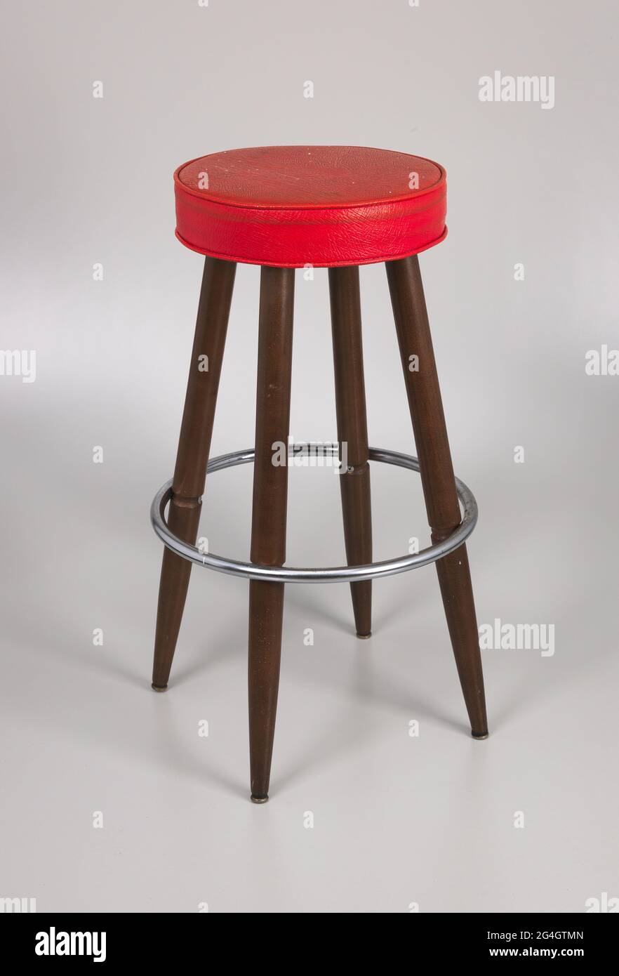 A wood barstool with a red vinyl seat. The seat of the stool is thick foam covered by red vinyl that is stapled to a particle board seat base. The stool has four legs made of lathe-shaped wood, with each leg tapering at the tip and finished with a metal cap. The legs bolt into the underside of the seat, so that the seat does not swivel. 1 foot up from the ground, a circle of metal is attached horizontally to the stool legs, which serves as a foot rest. The metal circle attaches to each leg by one long metal screw that runs through each leg and screws into the metal. Stock Photo