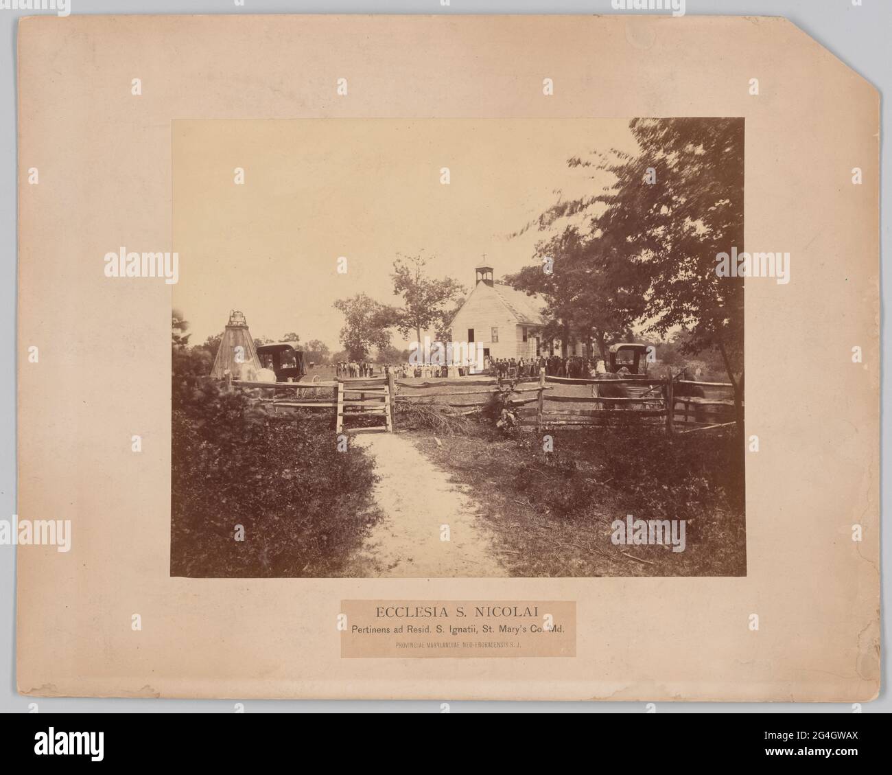 A reddish-brown photograph of the African-American congregation of St. Nicholas Church in St. Ignatius, St. Mary&#x2019;s County, Maryland. The image depicts a large group of people standing in front of Saint Nicholas Church. The foreground depicts a dirt path leading to a wooden fence. A white horse is near the fence on the left side of the photograph. Trees are on the right side of the photograph. Behind the fence is a large field with the church at center. A line of women and men are standing in front of the building. The women appear to be wearing white colored clothing and the men appear Stock Photo