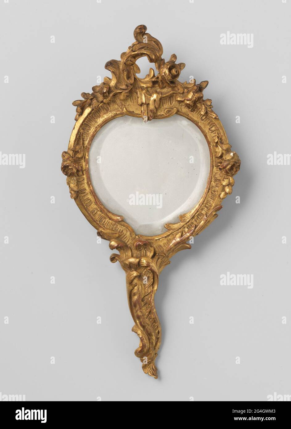 Hand mirror with handle, irregularly round of shape, with carving in C and S shapes and flowers and leafwork and crowned by C-shaped crest with stalactite with two roses .. Wooden cut and gold-plated hand mirror consisting of a handle and the organically connected Mirror frame, which is irregularly round in shape. The carving consists of stretched S and C forms, leafwork and flowers. The mirror is crowned in the middle by a C-shaped crest with a stalactite and two small roses. Small roses are also installed for decoration at halfway. The rear has probably been placed with red fabric; Traces st Stock Photo