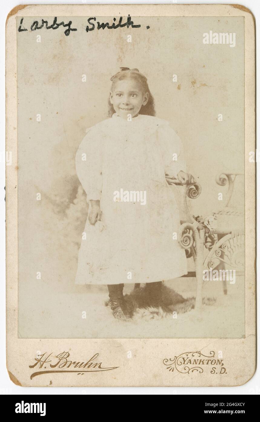 A black-and-white cabinet card photograph of a young African-American girl wearing a white dress. She has a dark colored ribbon in her hair and has her right hand resting on a wicker chair. She is smiling at the camera. Written in black ink at the top right corner of the photograph is &#x201c;Larby Smith.&#x201d; Printed on the cardstock below the photograph is &#x201c;H. Bruhn&#x201d; and &#x201c;Yankton, S. D.&#x201d; There are no inscriptions on the back. Stock Photo