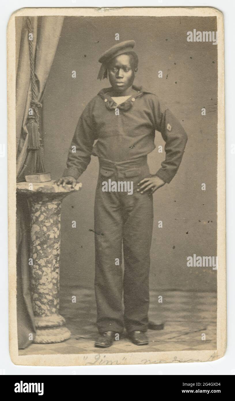An African American sailor standing with his right hand resting on a small pillar. His left hand is resting on his hip and his left arm is bent at the elbow. He is wearing a sailor&#x2019;s uniform with an unidentifiable lozenge on left sleeve. A book is on the pillar and a curtain is on the left side of the photograph. Below the image written in pencil is &#x201c;Jim&#x201d; and &#x201c;my servant.&#x201d; Stock Photo