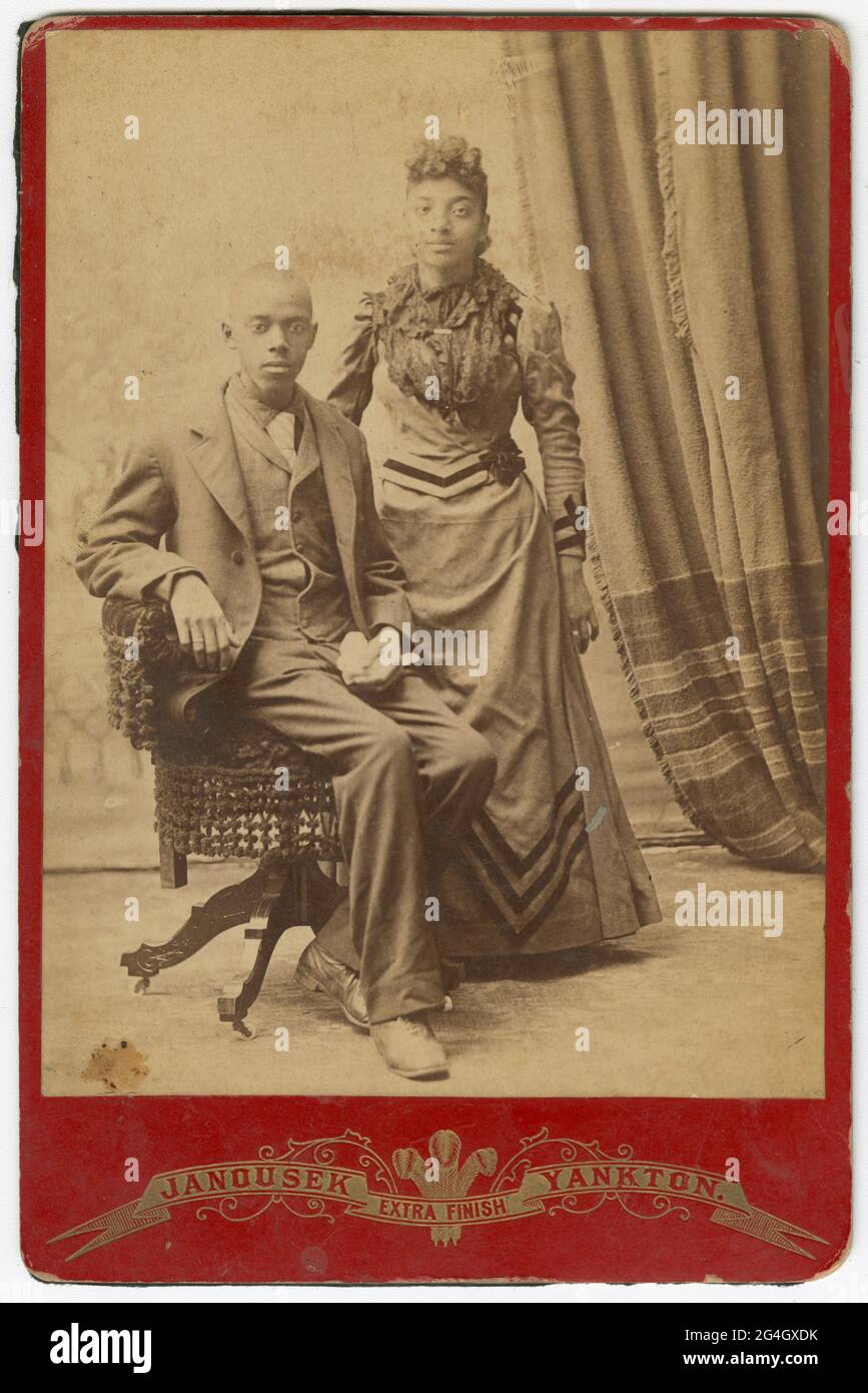 A black-and-white cabinet card photograph of a man seated on a wicker chair with a woman standing next to him. The man is wearing a dark colored suit and vest and a light colored necktie. He is holding a handkerchief in his left hand. The woman standing next to him is wearing dark colored dress with even darker stripes. A curtain is in the background on the right side of the photograph. The photograph is mounted on red cardstock. Below the photograph printed on the cardstock is &quot;Janousek,&quot; Extra Finish&quot; and &quot;Yankton.&quot; There are no inscriptions on the front or back. Stock Photo