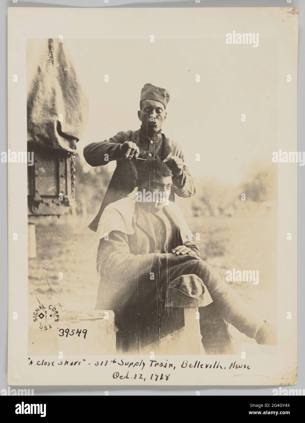 African-American soldiers serving in France during the First World War. One soldier is shaving another soldier's head. One soldier is seated with his right leg crossed over his left leg. His hands are resting on his thigh and he has a cigarette in his mouth. His hat is hanging on a wooden post to his right. The other soldier stands behind. His hands are raised and he is holding the razor blade in this right hand. He is wearing his uniform and hat and is pursing his lips. The Signal Corps USA logo is in the bottom left corner of photograph. Stock Photo