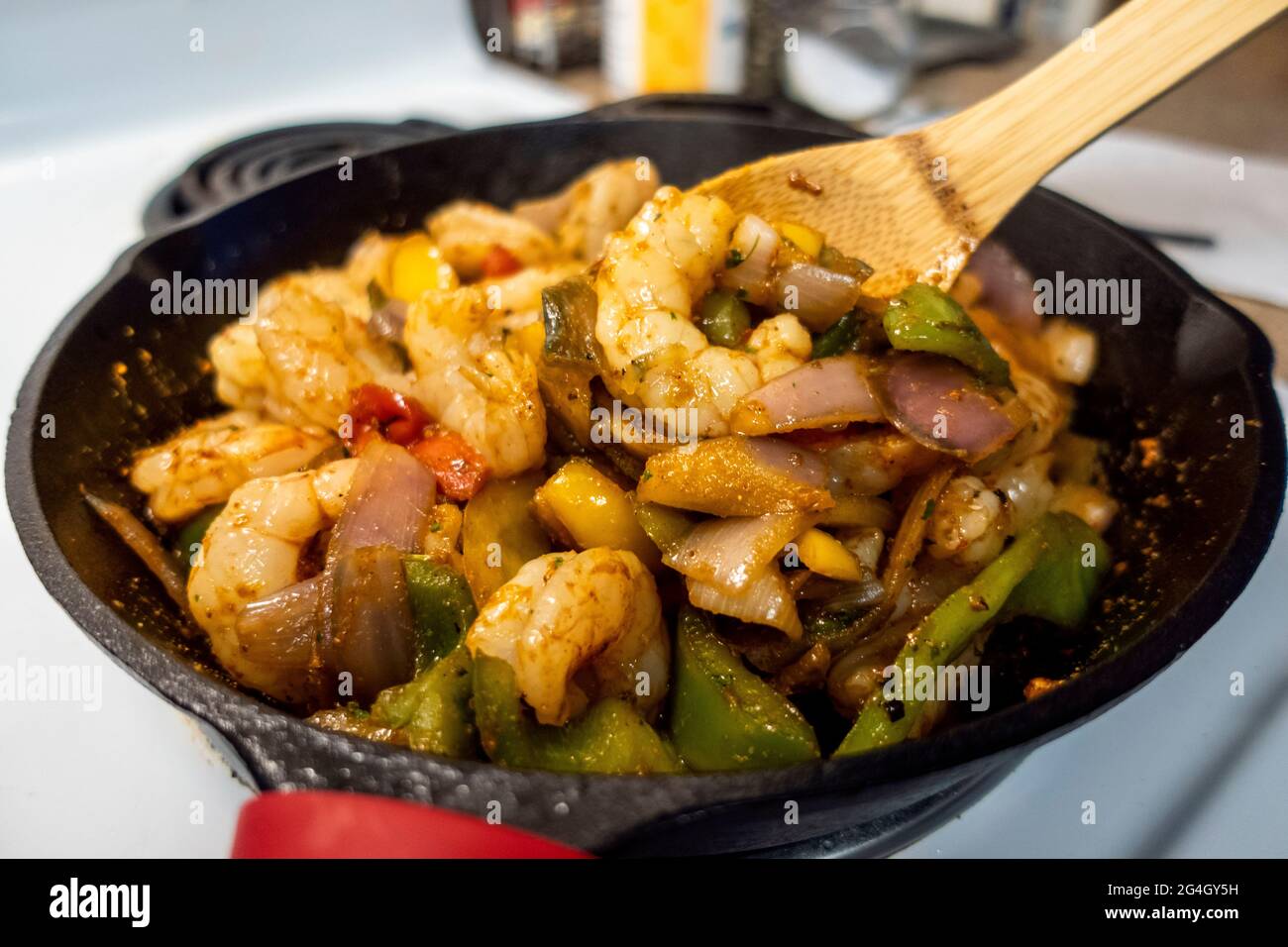 View of a wooden spoon stirring up shrimp and bell peppers, creating a fajita mixture in a cast iron skillet on a white stove top Stock Photo