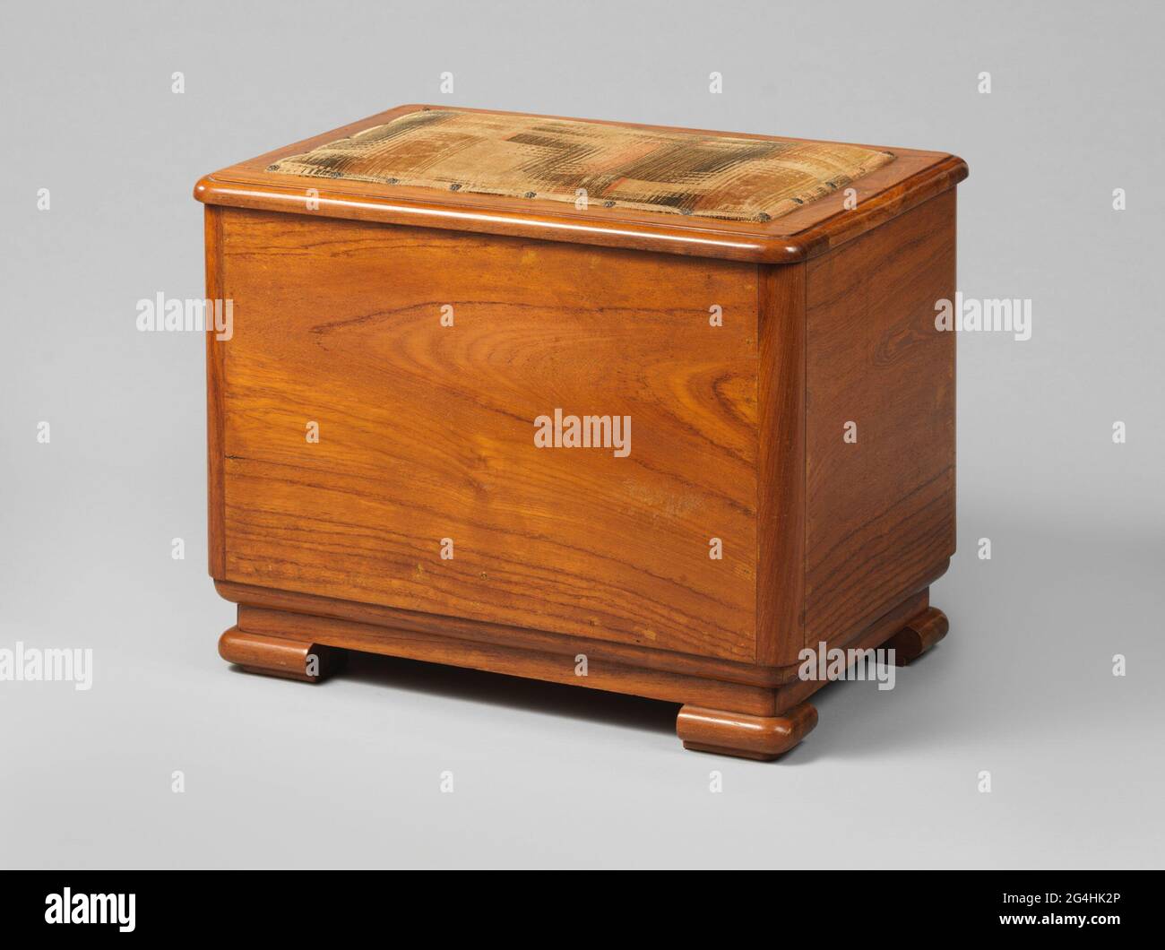 Chamber branch with lid lined with fabric. Chamometer of teak resting on four wide feet with rounded sides. The rectangular room branch has four walls whose rounded corners consist of one strip of wood. The lid, which is attached with two hinges, is coated with fabric woven with black, red and brown wires that is fixed with nails with heads in the shape of a flower. The inside is divided into two spaces whose right-hand side is greatest. The space on the left is closed at the height of the middle with a panel from which two circles are sawn. The space on the right is closed at the top with a p Stock Photo