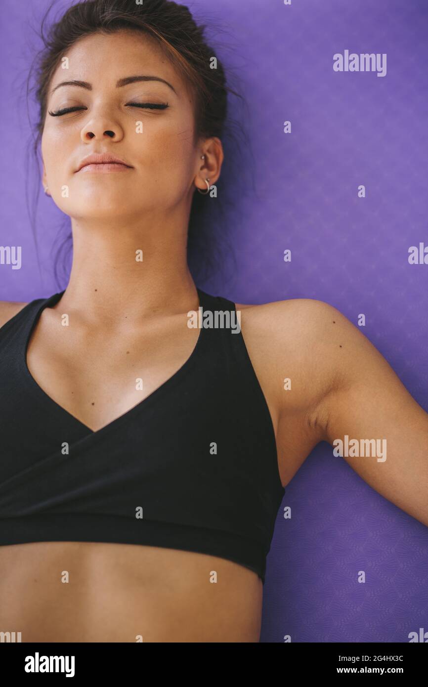 Female lying on a fitness mat with her eyes closed in meditation. Healthy young woman relaxing in savasana at gym. Stock Photo