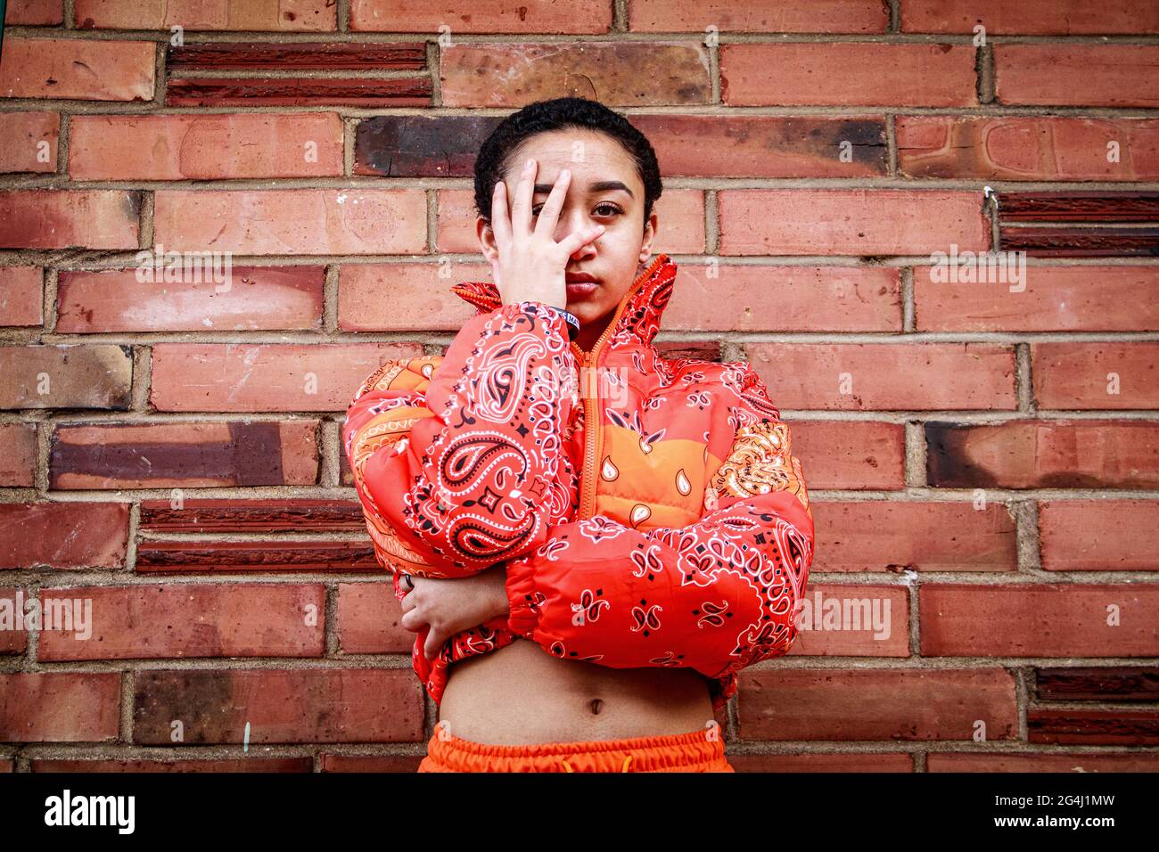 Titled: Power - Girl standing in front of brick wall, wearing an orange jacket. Stock Photo