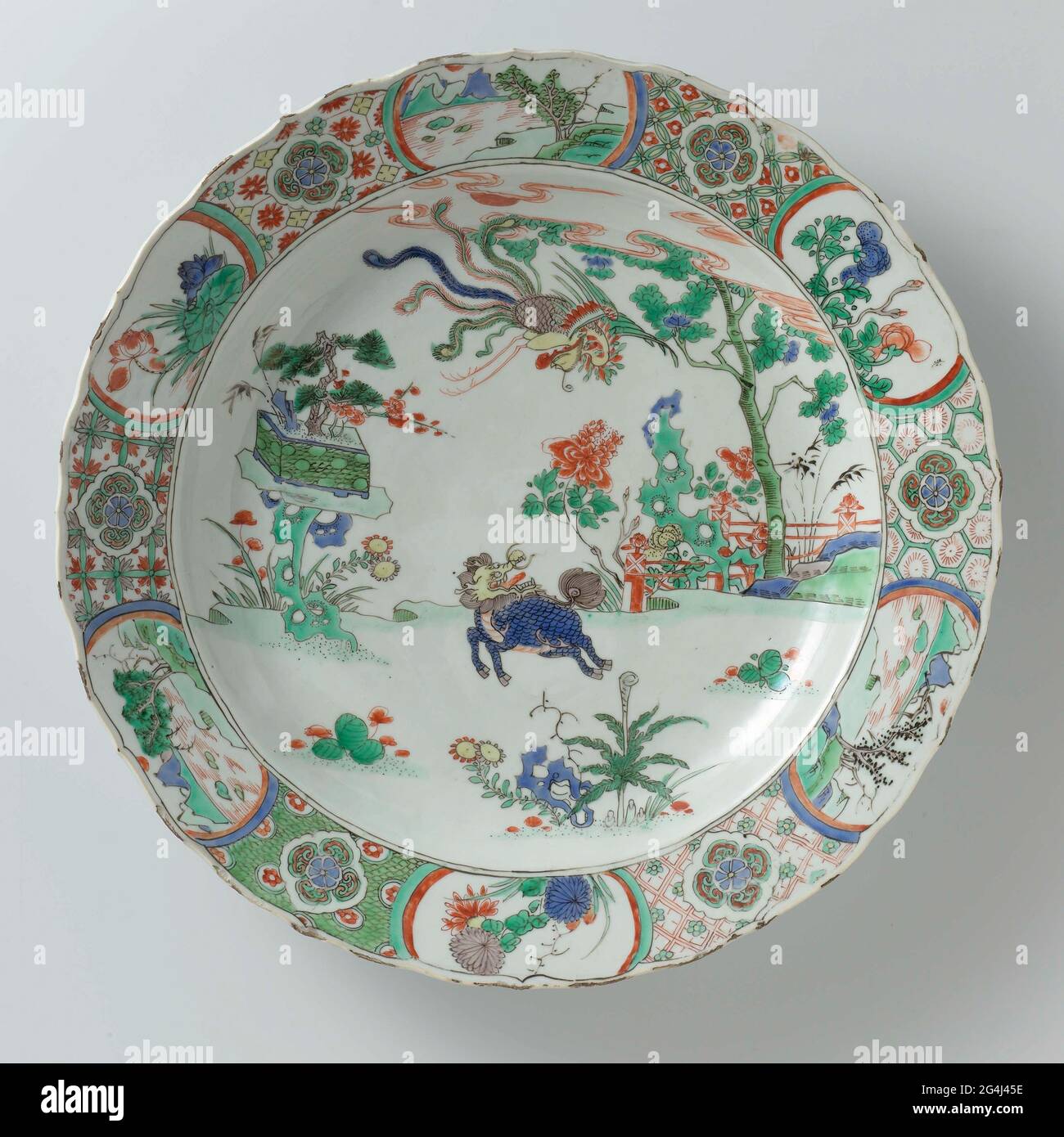 . Dish of porcelain with a scalloped edge, painted in underglaze blue and on the glaze with blue, red, green, yellow, eggplant and black. On the flat a Qilin and flying Feng Huang in a landscape with right rocks, peony plant and tree for a fence and left a pot with prunus, pine and bamboo on a rock; The edge with six times a flower rosette with four ruyi motifs in a lobd cartouches surrounded by a geometric pattern, interspersed by six times a plant (lotus, chrysanthemum, peony) or a river landscape in a cartouche. Marked on the bottom with an artemisia leaf in a double circle. Edge damaged. O Stock Photo