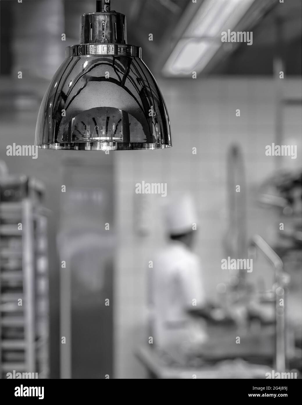Shiny, stainless warming light in foreground of a commercial kitchen with a chef wearing all white in the background. Stock Photo