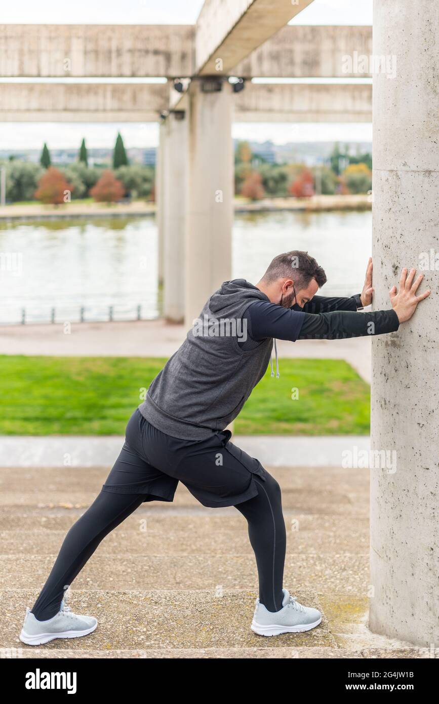 Runner with mask stretching in a park leaning against a wall. Stock Photo