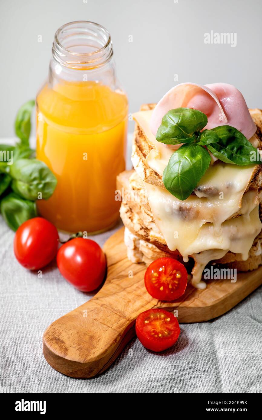 Stockpile of toasted melted cheese pressed sandwiches with ham meat, cherry tomatoes, orange juice and basil leaves on wooden cutting board on white m Stock Photo