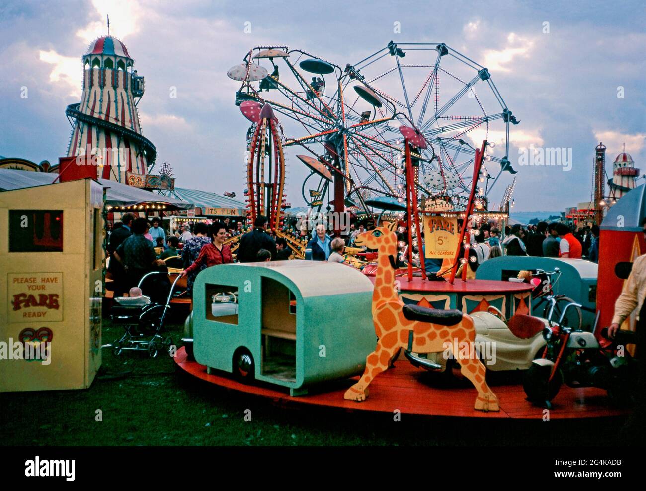 Crowds at dusk at the Bridgewater Fair, Bridgewater, Somerset, England, UK in September 1971. The rides include the big wheel, helter-skelter and various roundabouts and merry-go-rounds. The lights are on and traditional, colourful signs of hand-painted fairground lettering and art are visible. The St Matthews fair dates back to 1249. The traditional annual four-day event takes place in St Matthew’s Field in September – a vintage 1970s photograph. Stock Photo