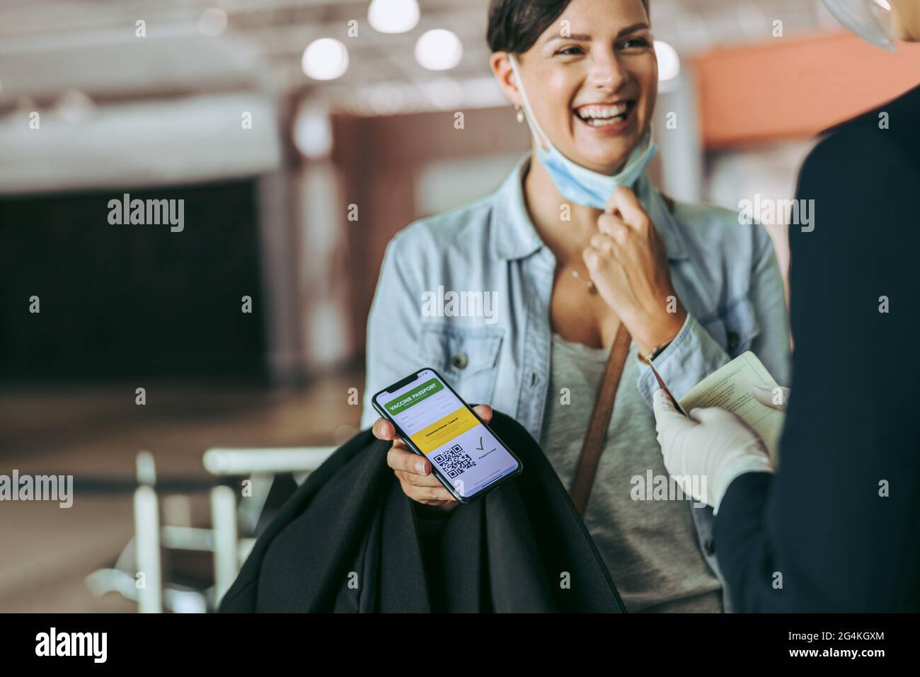 Smiling woman showing immunity passport to security at airport. Female at check-in counter showing her vaccine passport to the airline attendant. Stock Photo