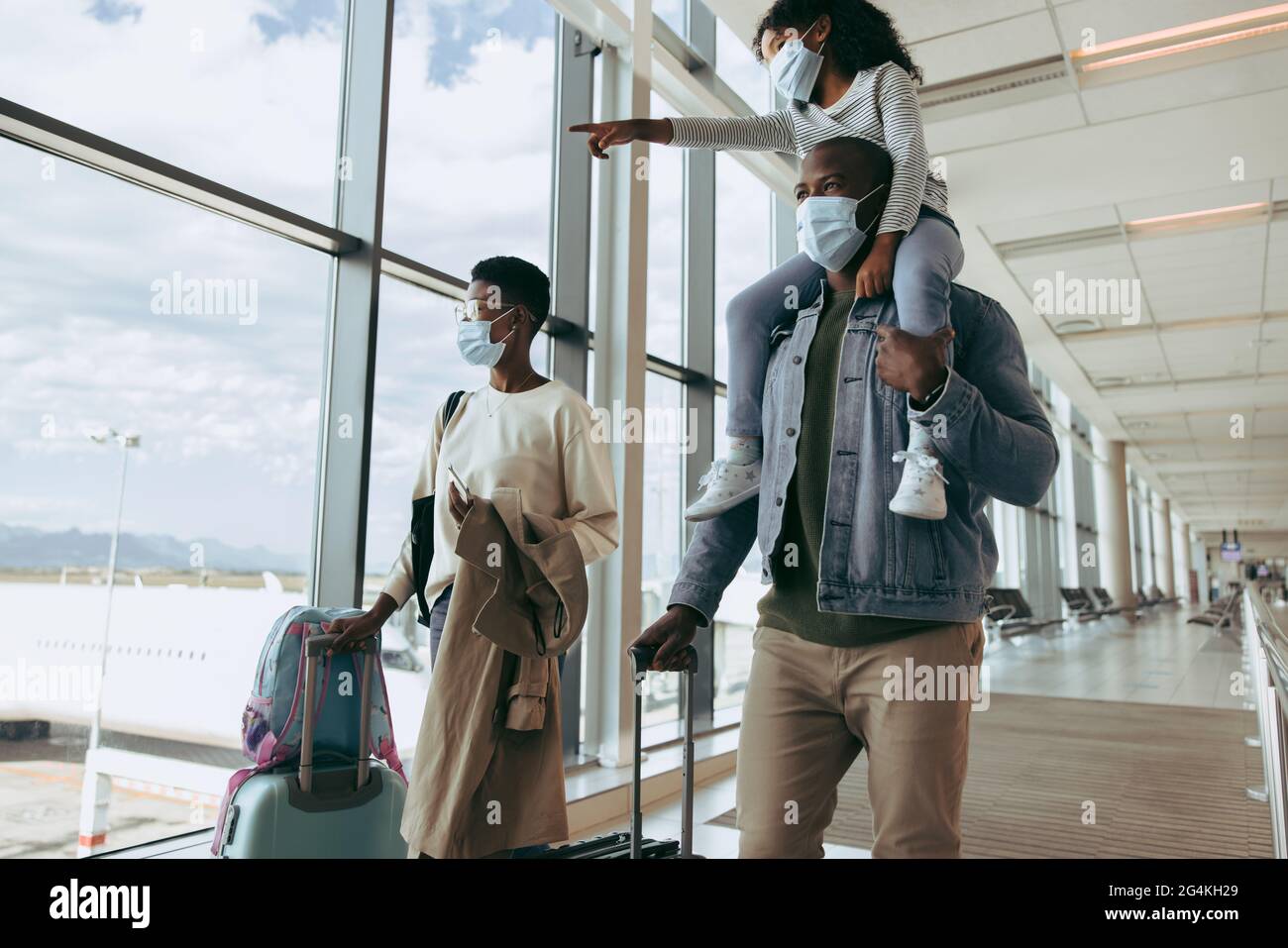 Kid girl on shoulder of man showing airplanes while walking at airport. Family wearing face masks going for boarding airplane. Stock Photo