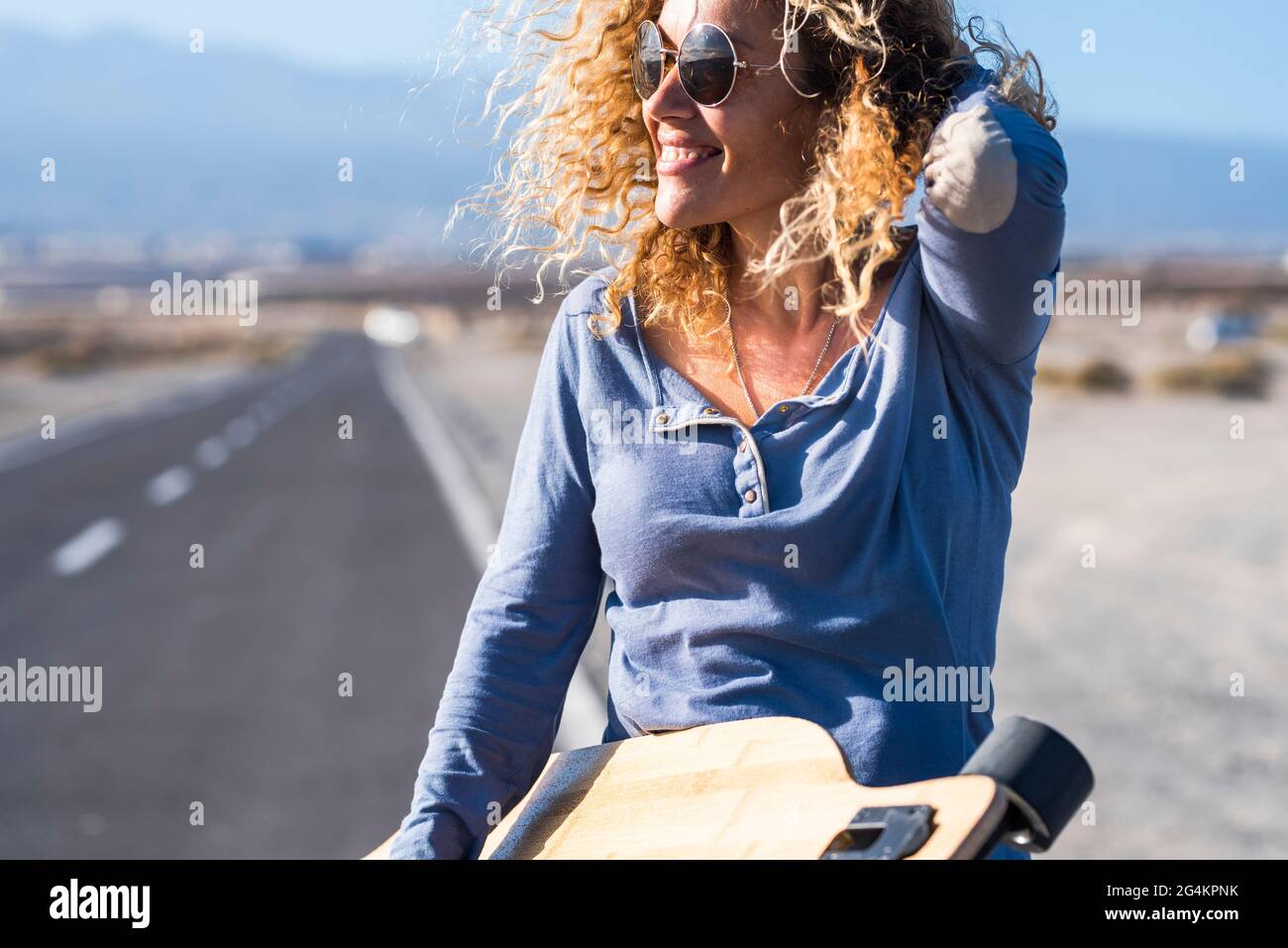 Cheerful attractive pretty adult woman smile and enjoy freedom using long skate board table with road in background - concept of travel and enjoy leiu Stock Photo