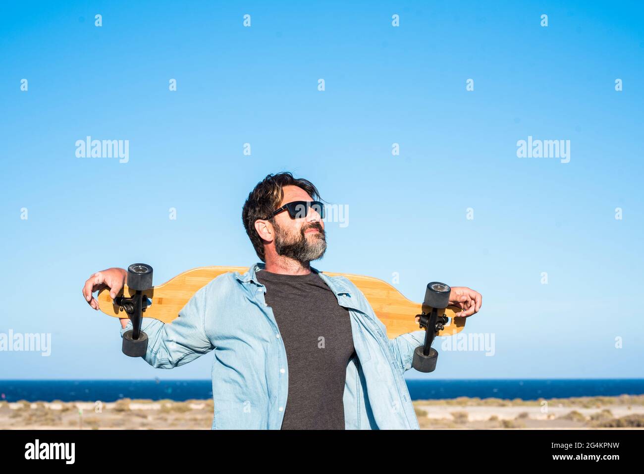 Standing free bearded handsome adult man with long board skate enjoying freedom and healthy lifestyle - blue ocean and sky in background - concept of Stock Photo