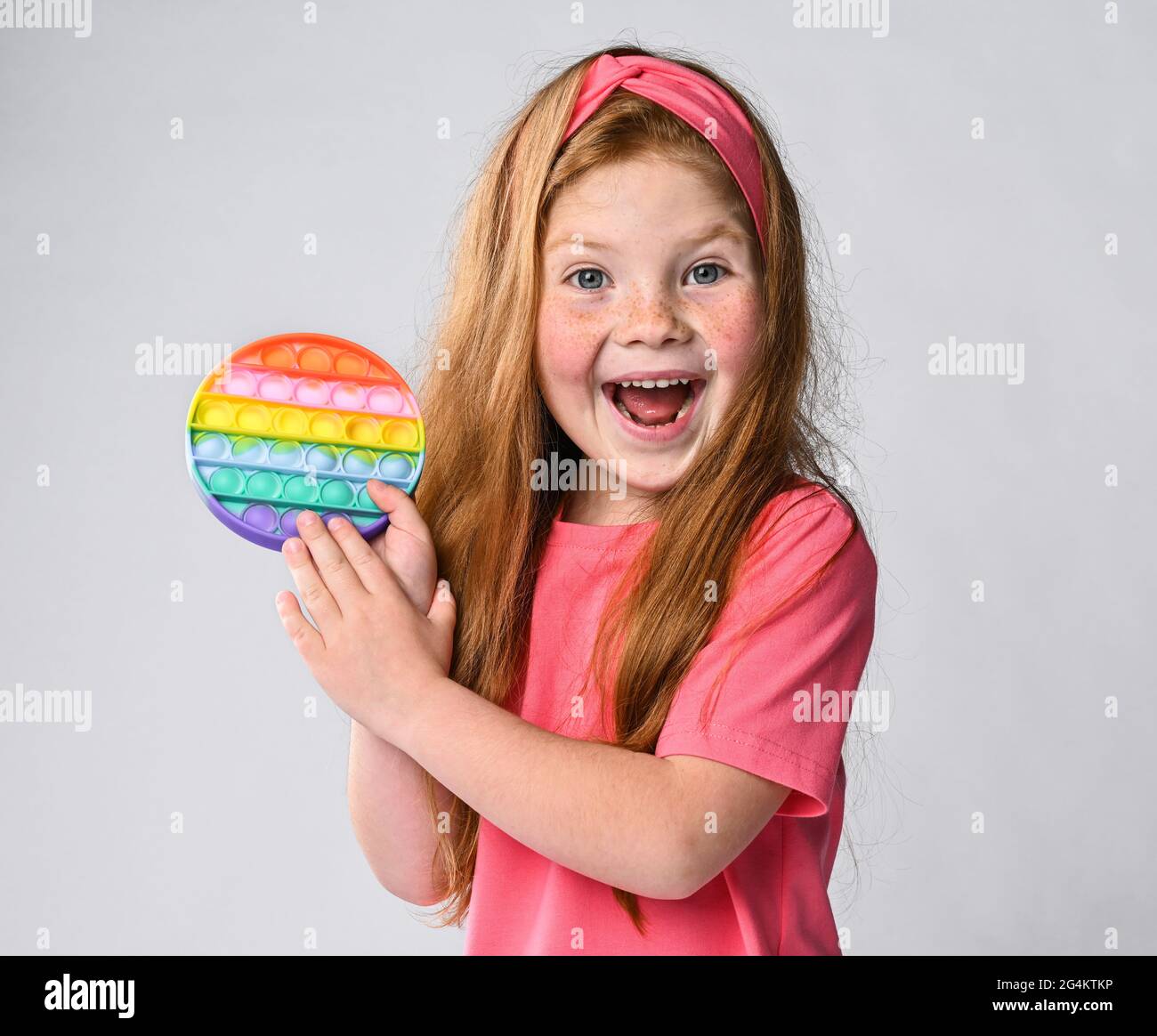 Portrait of happy screaming kid girl holding showing new sensory toy - pop it. Rainbow color, concave and convex bubbles Stock Photo