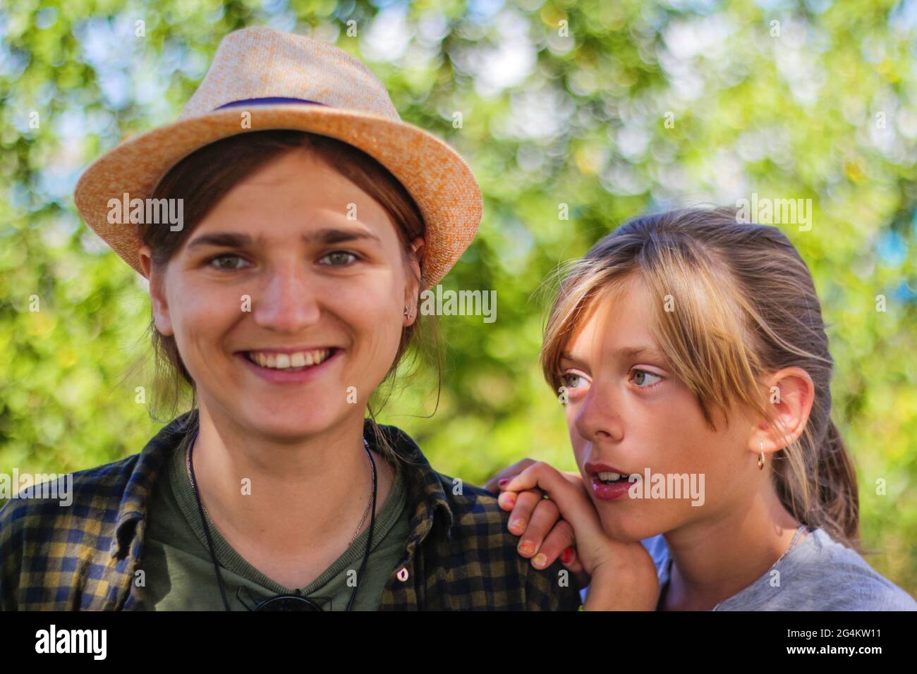 Defocused close-up smiling young brunette caucasian woman in hat with preteen girl. Summer blurred green nature bokeh background. Portrait of handsome Stock Photo