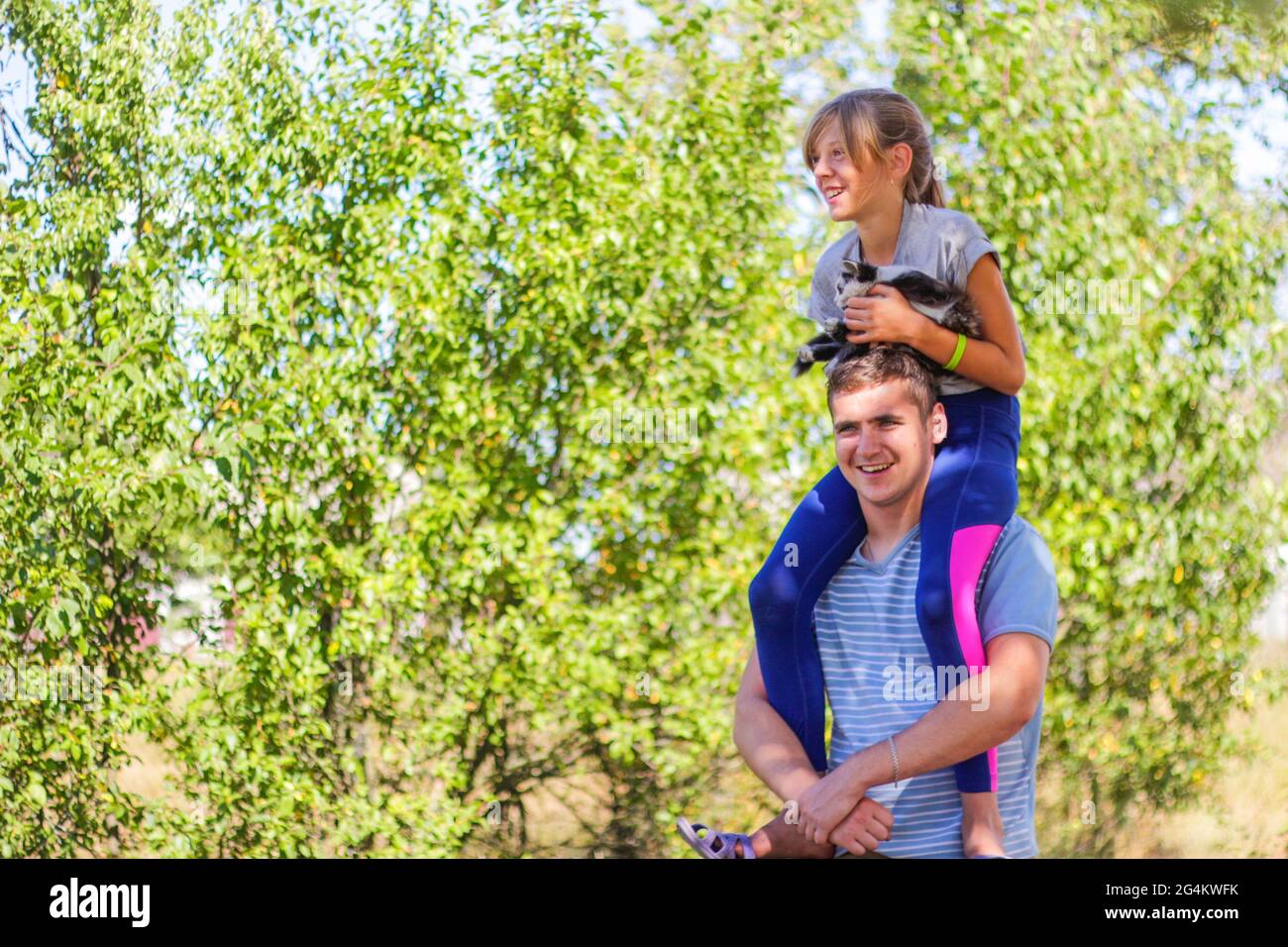 Defocused brother riding sister on back. Portrait of happy girl on man shoulders, piggyback. Girl holding cat, kitten. Family playing outdoor. Love an Stock Photo