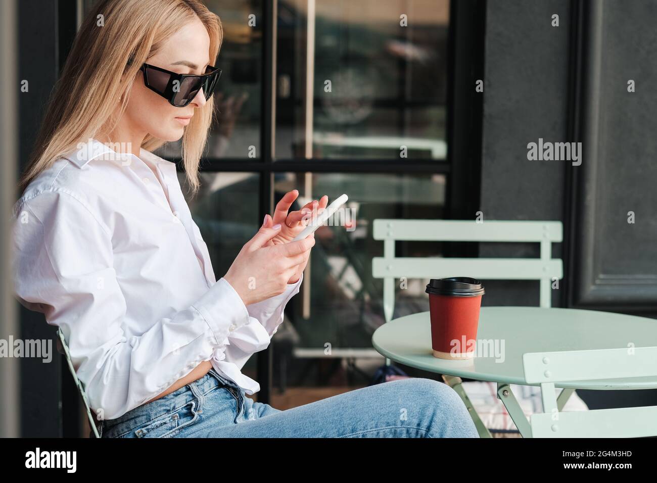 Side View of Elegant Caucasian Woman Sitting at a Cafe Table and Texting on Smartphone, Confident Female Entrepreneur Wearing Sunglasses and White Stock Photo