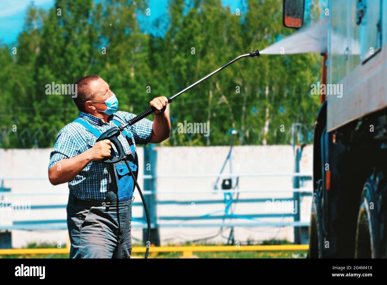 Car wash for special equipment and trucks. A male driver wearing a medical mask washes a truck with a pistol under pressure on the street on a summer day. Stock Photo