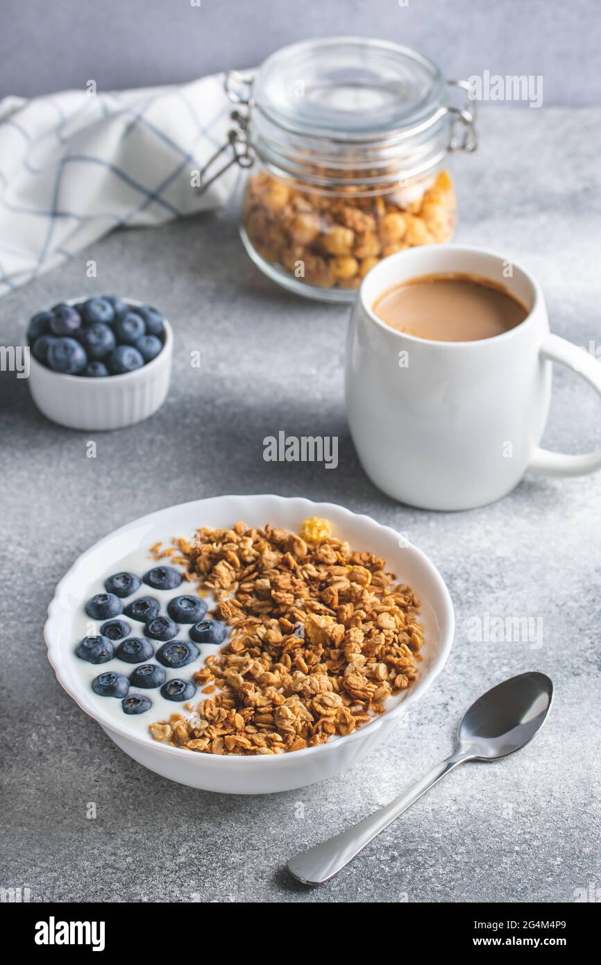Granola with yogurt and coffee on a gray table. Dry breakfast concept. Hot morning drink. Muesli and blueberries in a white bowl. Fitness diet. Health Stock Photo