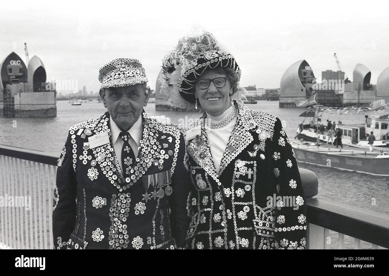 1980s, historical, London's Pearly King and Queen stand for their picture on the southern side of the river Thames at New Charlton, Greenwich, London, England, UK. Representatives of London's working class culture, the Pearlies, as they are known due to their clothes decorated with mother-of-pearl buttons, are standing in front of the newly constructed Thames Water Barrier, a retractable flood defence system. Operational since 1982, it was officially opened by Her Majesty Queen Elizabeth II in 1984. The barrier protects central London against a storm surge. Stock Photo