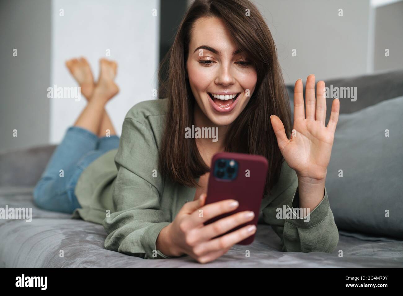 Young smiling woman gesturing and using mobile phone while lying on sofa at home Stock Photo